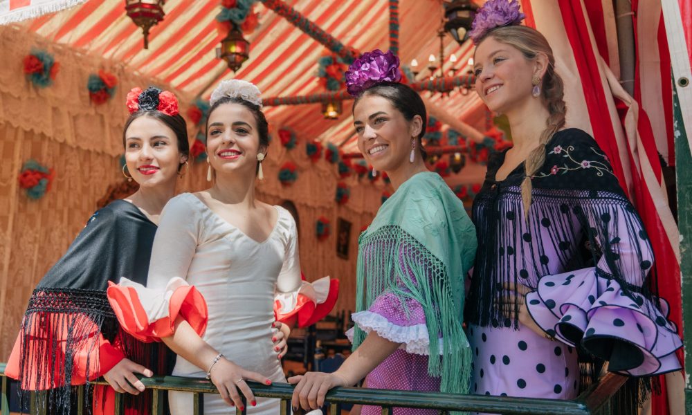 Group of smiling young women in colorful outfits standing near fence under striped tent with hanging decorations and looking away